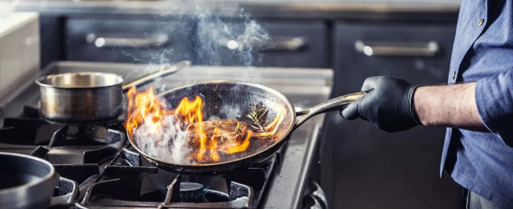 Chef cooking steak with flames in stainless steel frying pan on gas stove